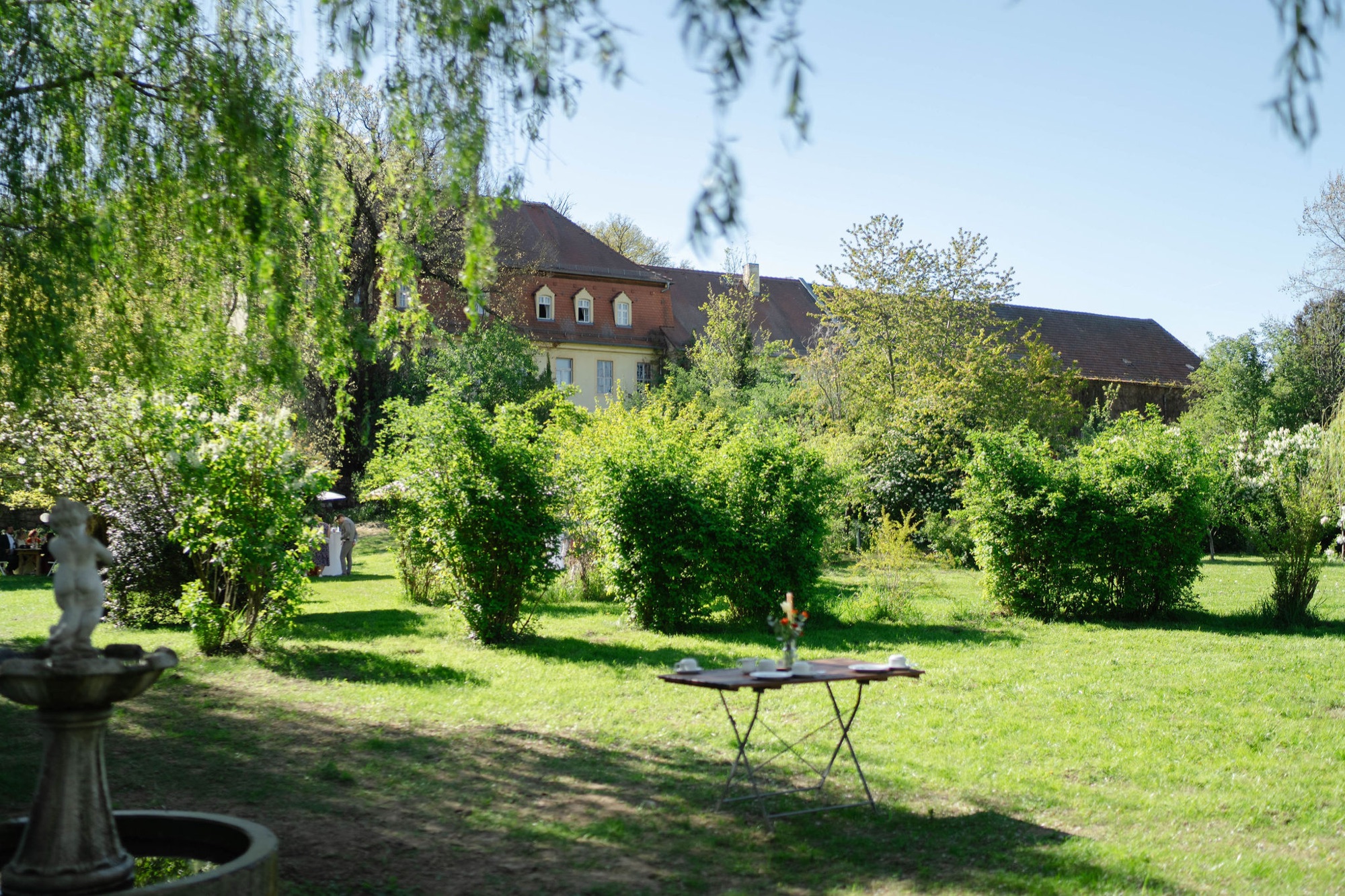 Der Garten mit Tisch und Stühlen unter Bäumen, dahinter das Gutshaus